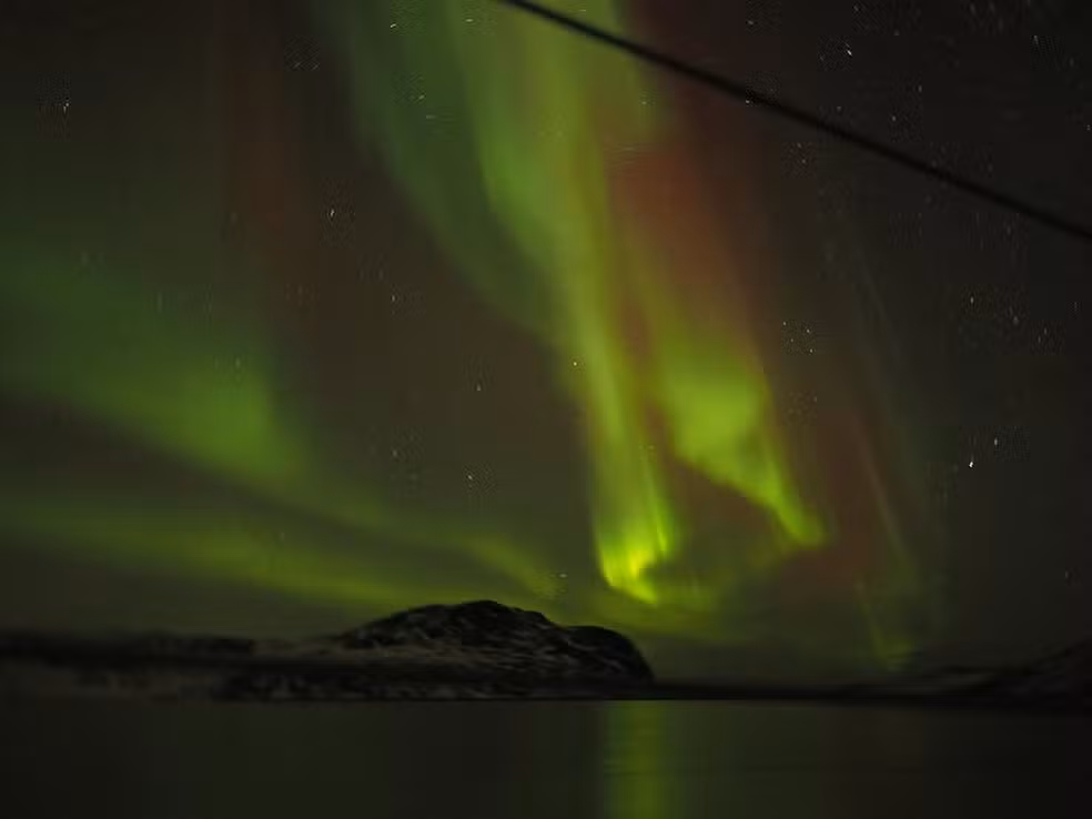 Aurora boreal vibrante com feixes de luz nas cores verde e avermelhada iluminando um céu noturno estrelado. Abaixo, a silhueta escura de montanhas e o reflexo suave das luzes na superfície da água ou gelo.