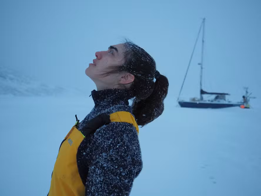 Perfil da navegadora Tamara Klink olhando para o céu enquanto flocos de neve caem sobre seu cabelo e rosto. Ela veste um casaco escuro e macacão amarelo de inverno. Ao fundo, uma paisagem branca e congelada com seu barco a vela parado no gelo.