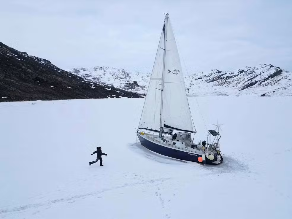 Tamara Klink, vestindo trajes escuros de inverno, caminhando sobre uma vasta superfície de gelo em direção ao seu veleiro, que está preso no mar congelado. Ao fundo, montanhas rochosas cobertas de neve sob um céu nublado.