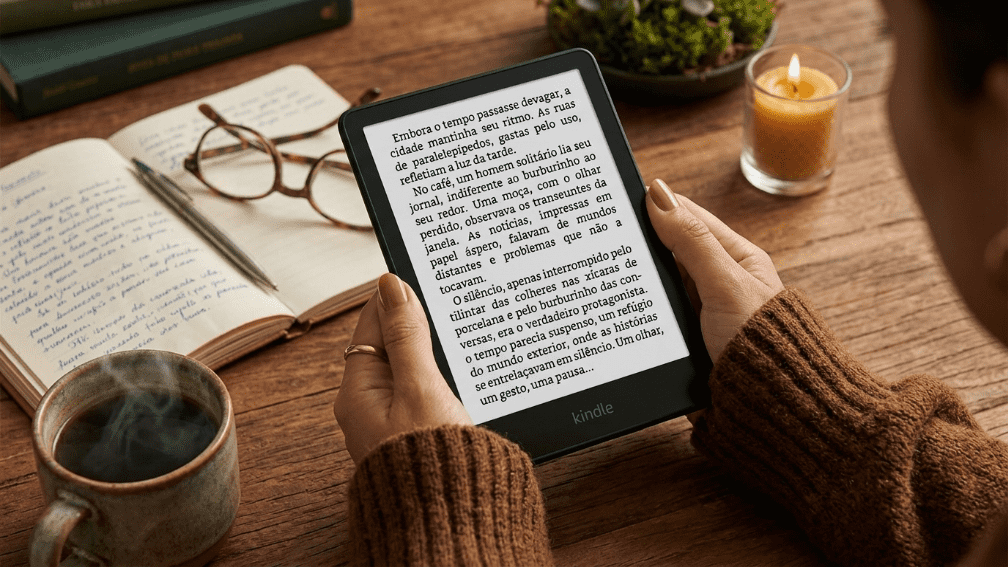Pessoa com suéter marrom segurando um leitor digital Kindle exibindo texto em português. O aparelho está sobre uma mesa de madeira aconchegante, acompanhado de uma xícara de café fumegante, um caderno aberto com caneta, óculos de grau e uma vela acesa.