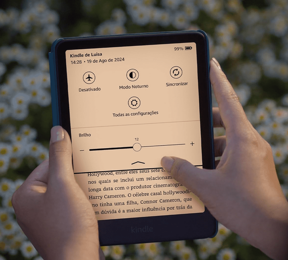 Mãos segurando um Kindle Paperwhite com capa azulada, cuja tela de tom quente (âmbar) exibe o painel de configurações rápidas de brilho e luz sobre a página de um livro. Ao fundo, um campo desfocado de flores brancas.